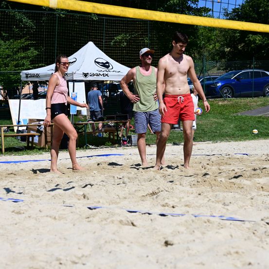 Drei Personen auf einem Beachvolleyballfeld, eine mit einem Ball. Zwei Männer stehen am Netz, während eine Frau sich nähert.