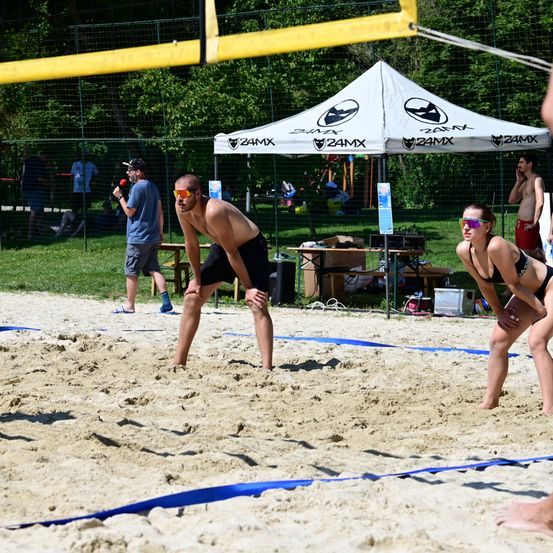 Eine Gruppe von Menschen spielt Beachvolleyball. Ein Mann beugt sich auf dem Sand. Eine Frau in einem schwarzen Bikini steht in der Nähe des Netzes.
