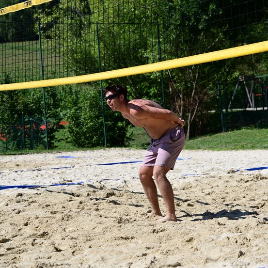 Ein Mann in Sonnenbrille spielt Beachvolleyball auf einem sandigen Platz und schaut zum Netz.