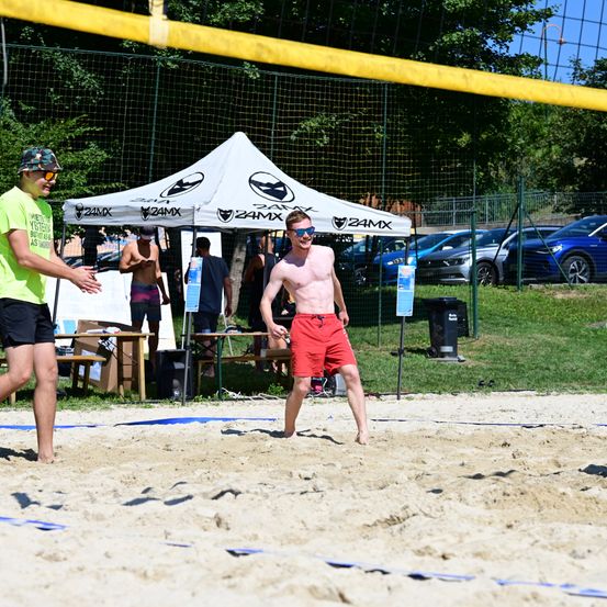 Zwei Männer spielen Beachvolleyball. Ein Mann ist ohne Hemd und trägt eine Sonnenbrille, während der andere in einem grünen T-Shirt ist. Dahinter steht ein Zelt mit einem Baldachin und sitzende Personen. Im Hintergrund sind Autos geparkt.