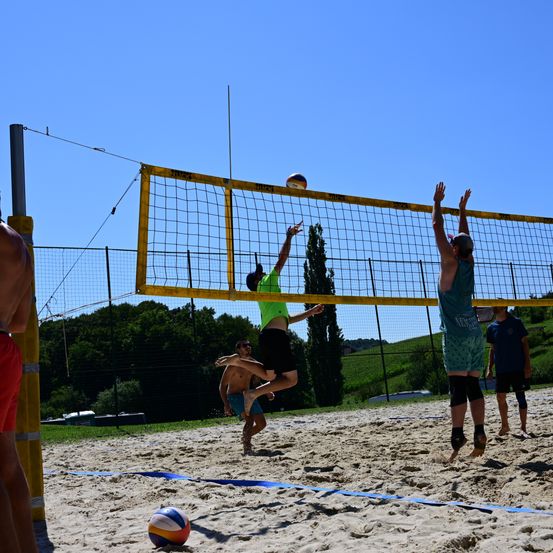 Zwei Männer springen, um einen Volleyball über das Netz zu schlagen, während ein anderer Mann zuschaut. Ein blau-weißer Ball liegt auf dem Sand.