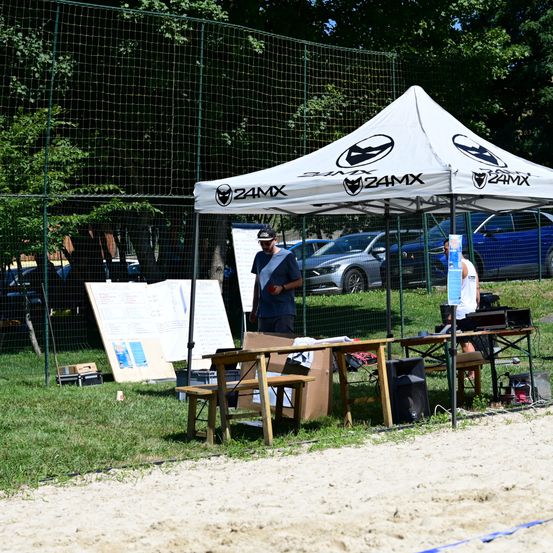 Zwei Personen stehen unter einem weißen Zeltdach in einem Grasbereich. Mehrere Autos sind im Hintergrund geparkt. Ein Mann in einer Kappe und einer Uhr steht in der Nähe des Zeltes. Eine andere Person sitzt auf einem Stuhl. Es gibt mehrere Schreibtische und eine Tafel auf dem Rasen.