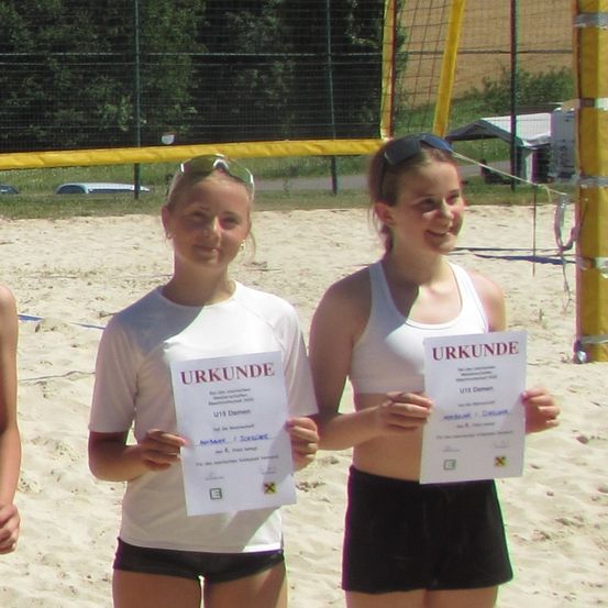 Zwei Frauen lächeln und posieren für ein Foto am Strand, während sie ein Zertifikat in den Händen halten. Sie tragen Sportkleidung und Sonnenbrillen. Hinter ihnen befindet sich ein gelbes Netz und ein Volleyballplatz.