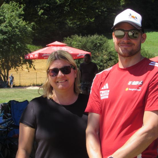 Ein Mann und eine Frau posieren für ein Foto im Freien. Die Frau trägt eine Sonnenbrille und ein schwarzes T-Shirt. Der Mann trägt ein rotes T-Shirt, eine Kappe und eine Sonnenbrille. Hinter ihnen befinden sich Bäume und ein Regenschirm.