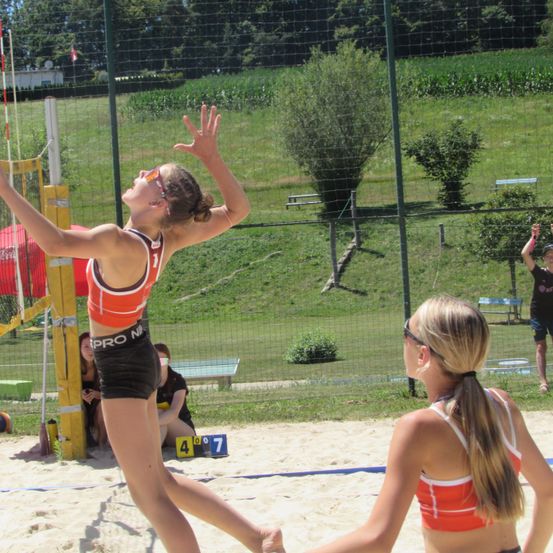 Zwei Frauen spielen Volleyball auf einem Sandplatz. Eine Spielerin springt, um den Ball zu schlagen, während eine andere zuschaut. Das Feld ist eingezäunt mit einem Netz und hat die Nummern 4 und 7.