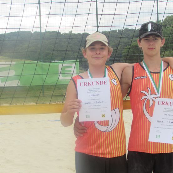 Zwei Jungen in orangefarbenen und gelben Trikots mit Medaillen posieren mit Zertifikaten in einer Beachvolleyball-Umgebung.