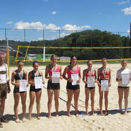 Eine Gruppe junger Frauen posiert am Strand mit ihren Urkunden und Medaillen. Ein Volleyballnetz ist im Hintergrund.