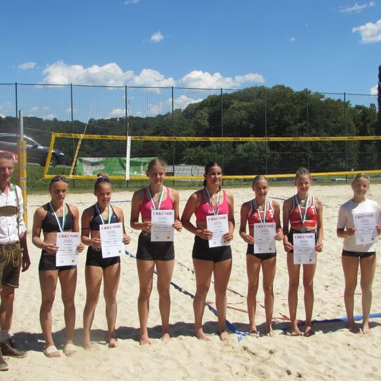 Eine Gruppe junger Frauen posiert am Strand mit ihren Urkunden und Medaillen. Ein Volleyballnetz ist im Hintergrund.