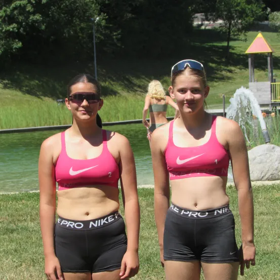 Zwei Frauen in pinkfarbenen Nike-Sport-BHs und schwarzen Shorts stehen neben einem Wasserbrunnen. Die Frau auf der rechten Seite trägt eine Sonnenbrille auf dem Kopf. Hinter ihnen geht eine Frau in einem grünen Bikini in der Nähe des Wassers. Bäume und Gras sind im Hintergrund.