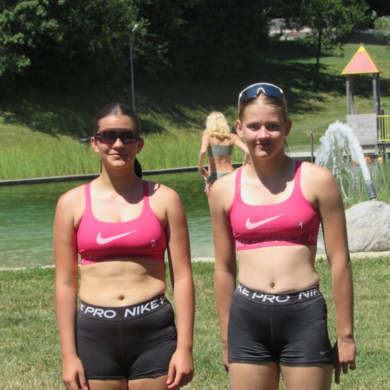 Zwei Frauen in pinkfarbenen Nike-Sport-BHs und schwarzen Shorts stehen neben einem Wasserbrunnen. Die Frau auf der rechten Seite trägt eine Sonnenbrille auf dem Kopf. Hinter ihnen geht eine Frau in einem grünen Bikini in der Nähe des Wassers. Bäume und Gras sind im Hintergrund.