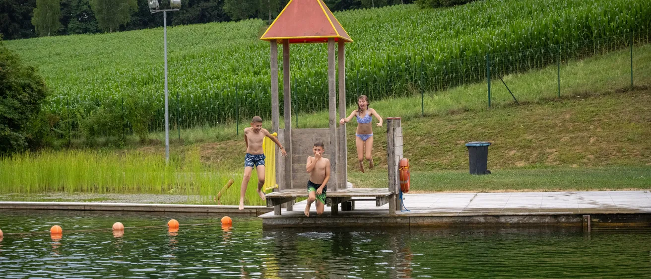 Drei Kinder springen von einer kleinen Plattform in einen See in einer ländlichen Gegend. Dahinter befindet sich ein Maisfeld.