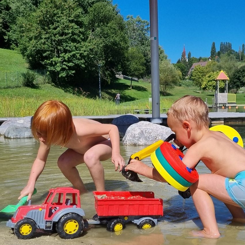 Zwei kleine Jungen spielen im Wasser mit Spielzeuglastwagen und Schaufeln in einem Wasserspielplatz.
