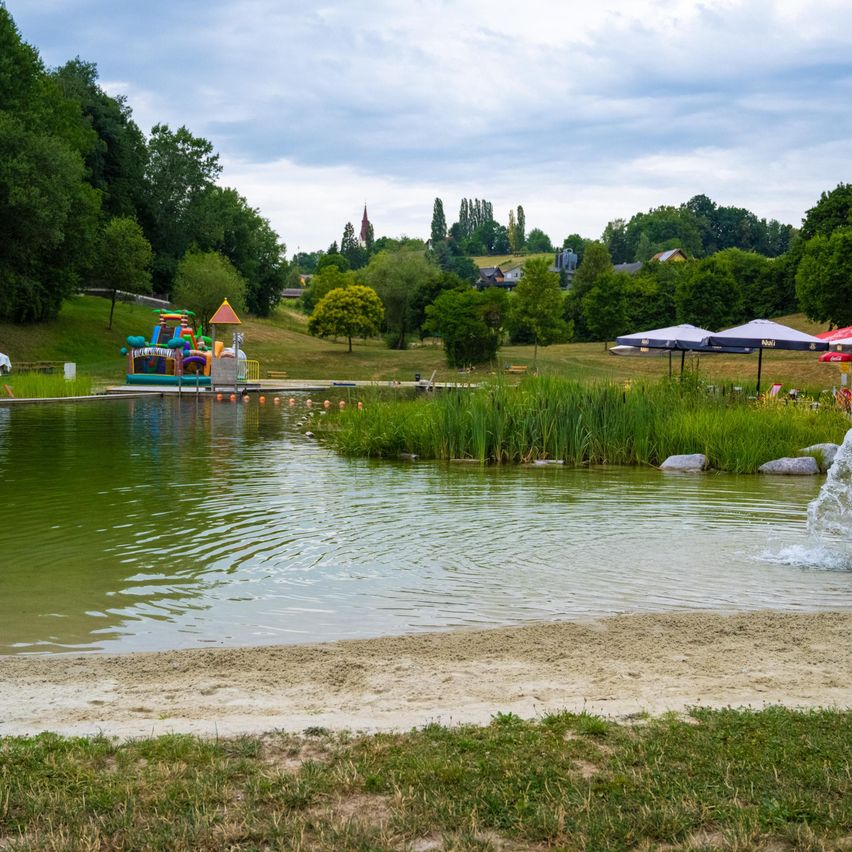 Ein Wasserpark mit einem bunten aufblasbaren Schloss, umgeben von Bäumen und Gras. Der See ist ruhig mit einem Springbrunnen. In der Ferne gibt es Häuser und einen Kirchturm.