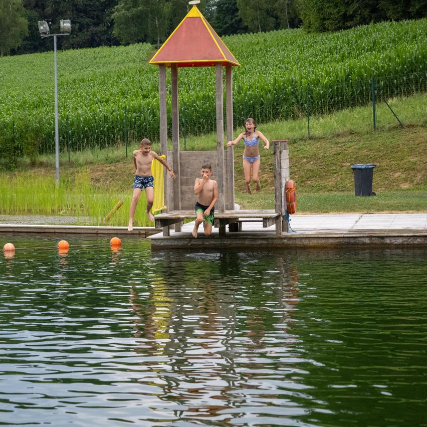 Drei Kinder springen von einer kleinen Plattform in ein Becken in der Nähe eines Pavillons, mit einem Maisfeld und einem grasbewachsenen Hügel im Hintergrund.