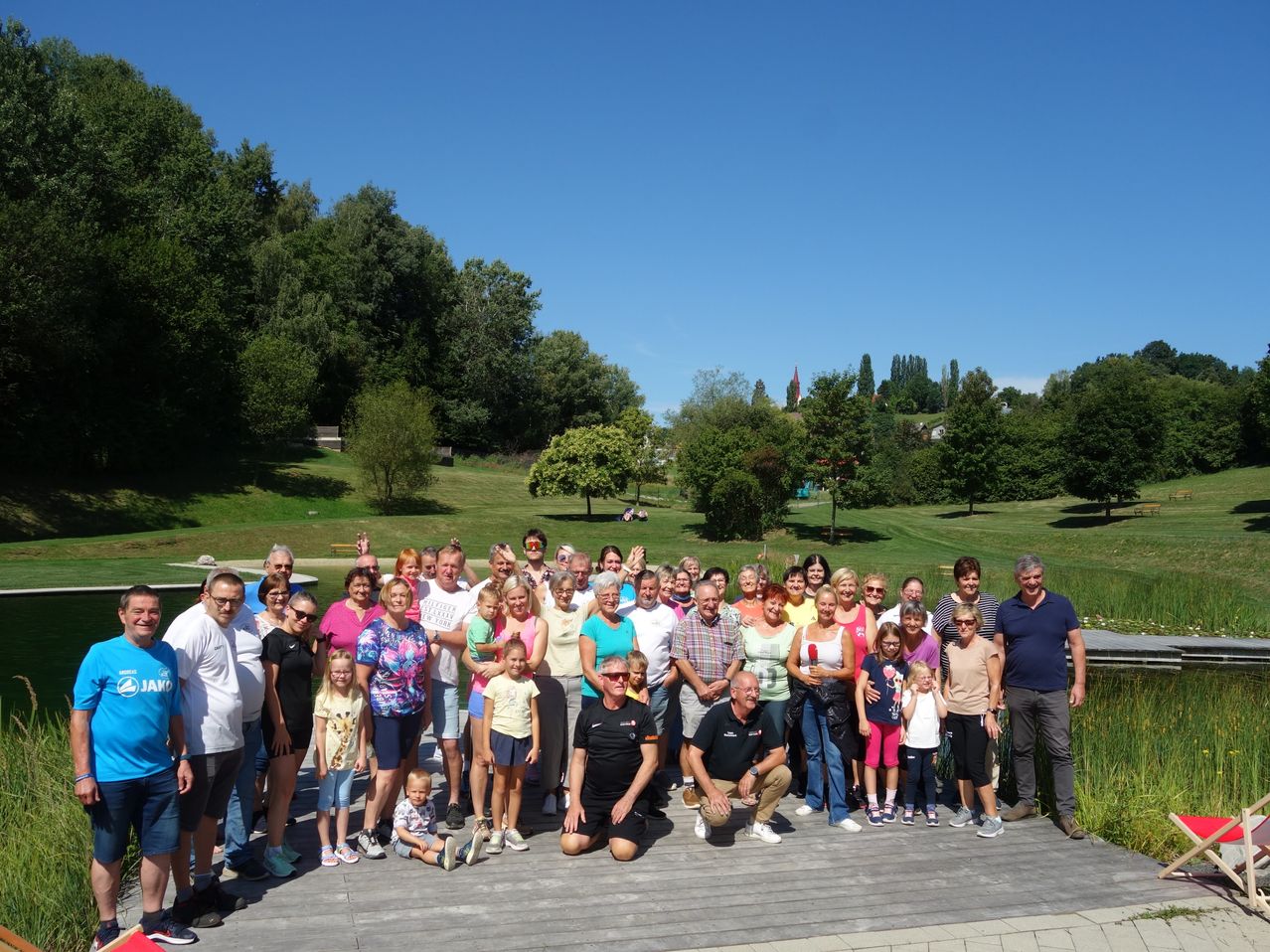 Eine Gruppe von Menschen posiert für ein Foto auf einer Holzterrasse in einem Park mit üppiger Grünfläche und klarem blauem Himmel. Einige tragen Sonnenbrillen, und ein Mann in einem schwarzen Shirt hockt vorne.