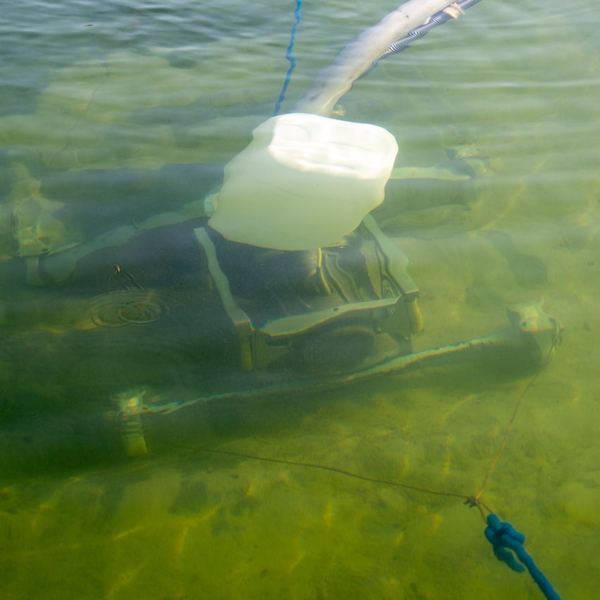 An underwater vehicle with a large white tank attached, submerged in green water with a blue rope connected.