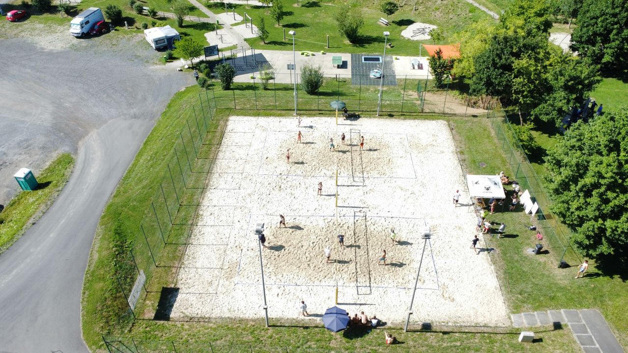 Ein großer Luftbild von einem sandigen Volleyballplatz, umgeben von einem Zaun. Mehrere Spieler spielen im Sand. Bäume und Bänke sind im Hintergrund.