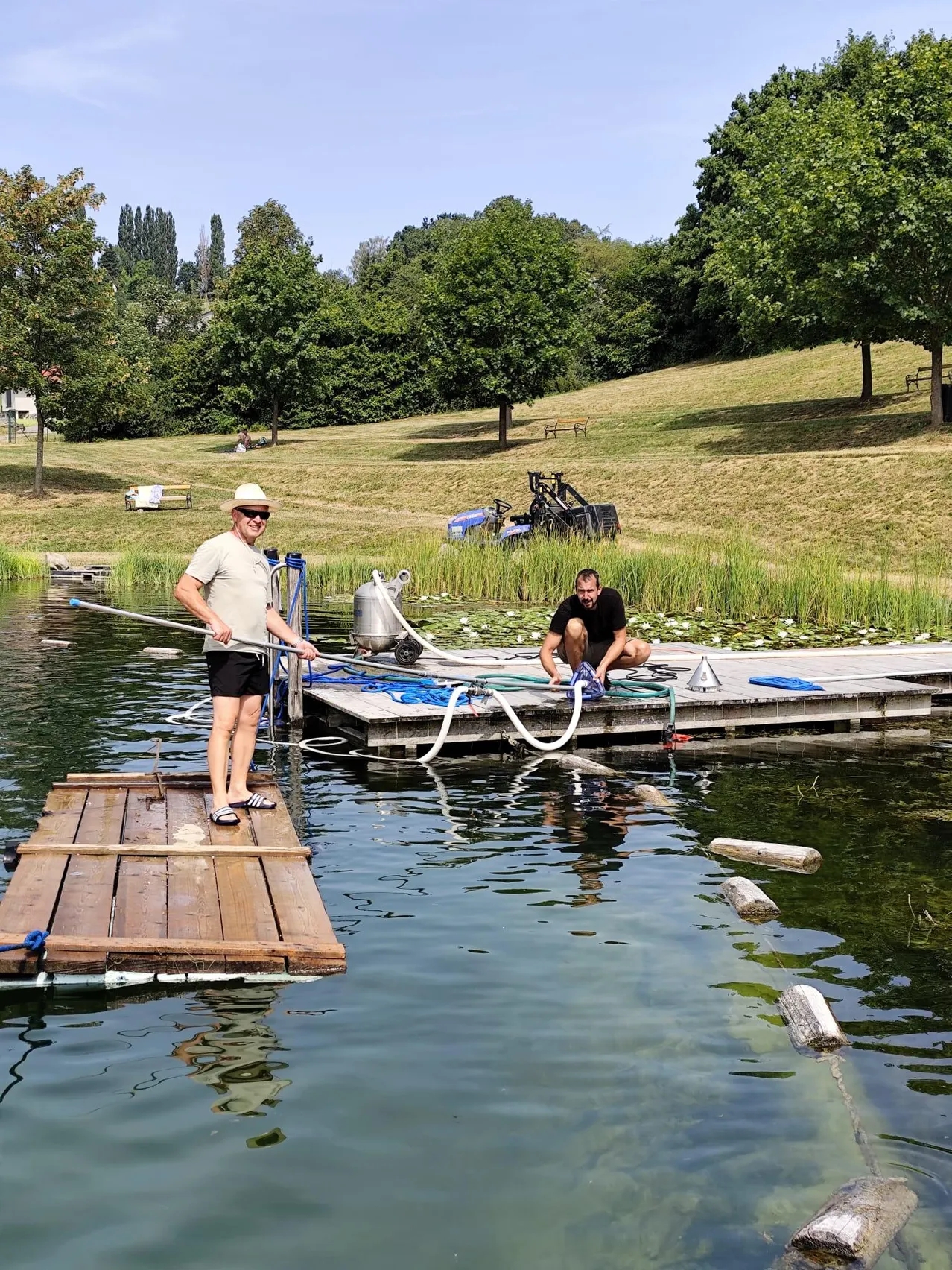 Zwei Männer arbeiten auf einer hölzernen Plattform in einem See, einer steht und hält einen langen Stab, der andere kniet und repariert etwas. Der See ist von Grün und einem grasbewachsenen Hügel umgeben.