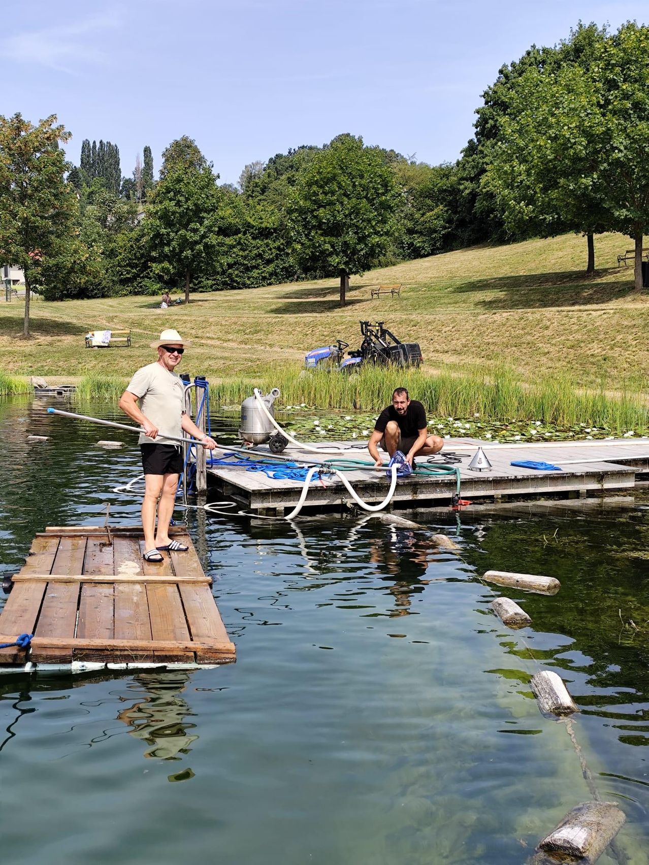 Two men are working on a wooden raft in a lake, one standing and holding a long pole, the other kneeling and fixing something. The lake is surrounded by greenery and a grassy hill.