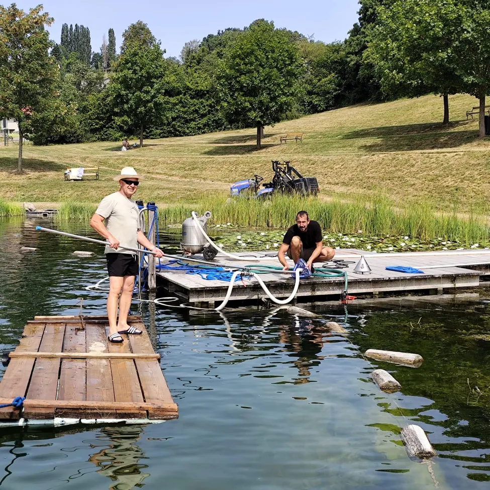 Zwei Männer arbeiten auf einer hölzernen Plattform in einem See, einer steht und hält einen langen Stab, der andere kniet und repariert etwas. Der See ist von Grün und einem grasbewachsenen Hügel umgeben.