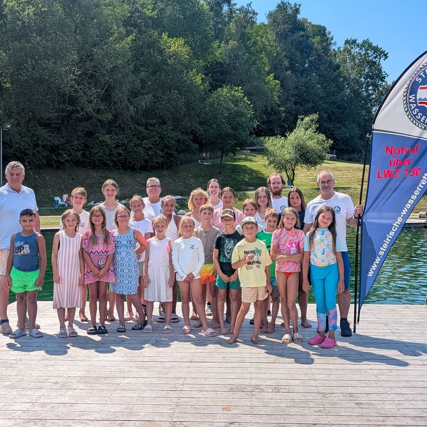Eine Gruppe von Kindern und Erwachsenen steht auf einem Holzsteg am Schwimmbad, mit einem blauen Banner im Hintergrund.
