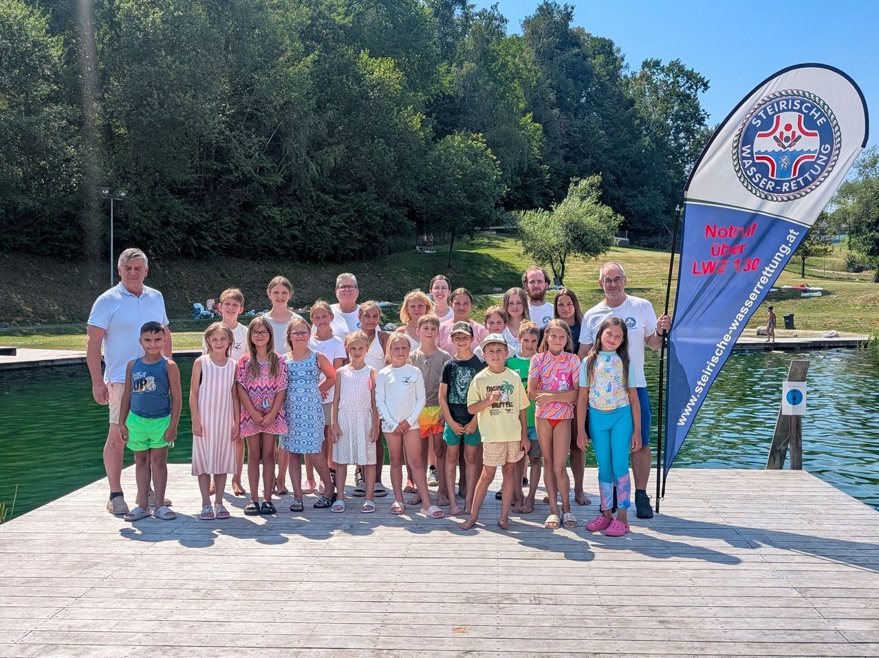 A group of children and adults stand on a wooden dock by a swimming pool, with a blue banner in the background.