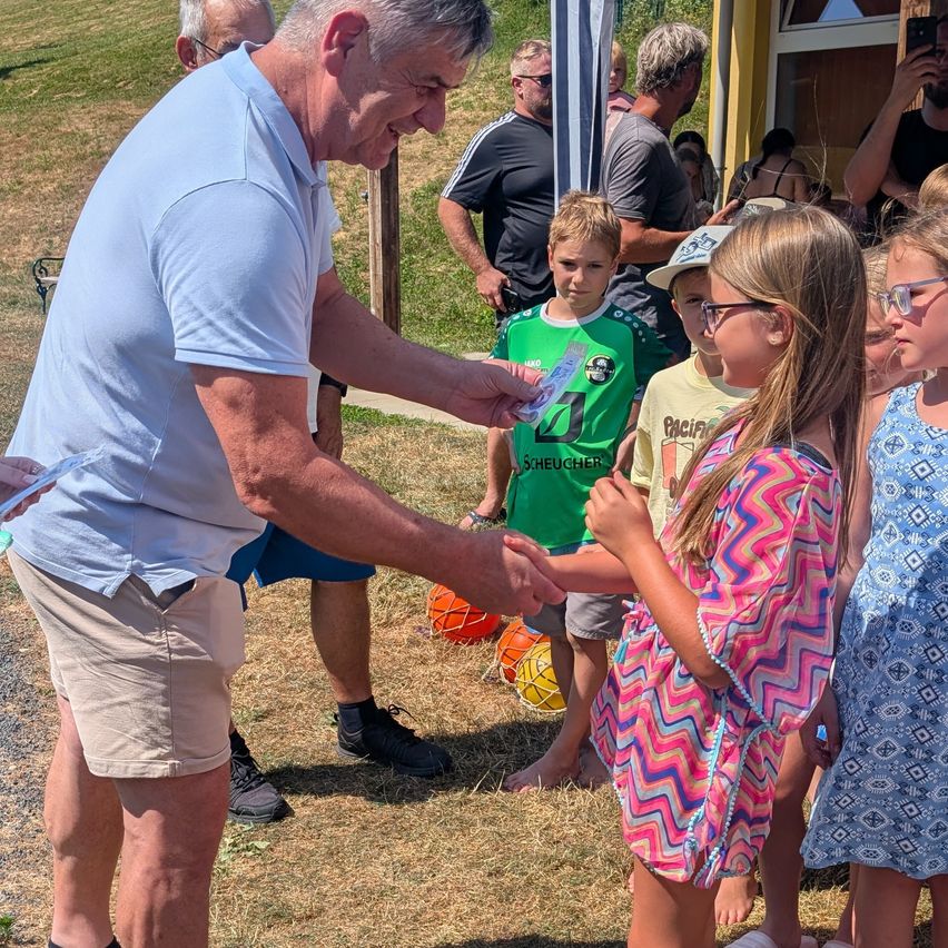 An older man hands a prize to a young girl with glasses, surrounded by other children and adults on a grassy field.
