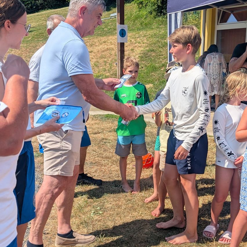 A man hands a certificate to a young boy in a white shirt with graphics, surrounded by other children and adults in an outdoor setting.