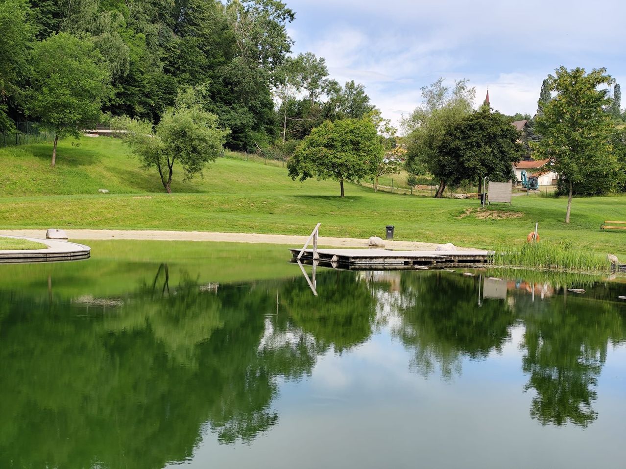Ein ruhiger See mit einem hölzernen Steg und üppigen grünen Bäumen, die den Himmel und Wolken an einem klaren Tag widerspiegeln.