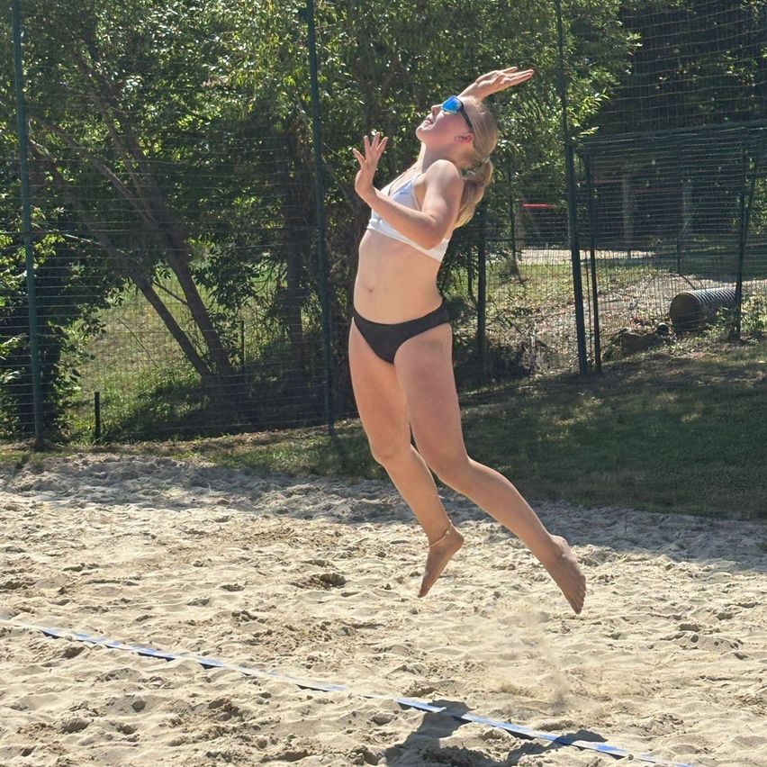 Eine Frau in einem weißen Bikini springt mit erhobenen Armen auf einem Sandplatz, wahrscheinlich beim Beachvolleyball spielen.