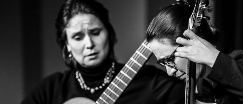 Ein Schwarz-Weiß-Bild zeigt zwei Frauen, die Gitarre spielen. Eine Frau trägt eine Brille und konzentriert sich auf ihr Instrument, während die andere Frau hinsieht. Beide haben ihre Hände auf den Gitarren, was auf eine musikalische Aufführung hindeutet.