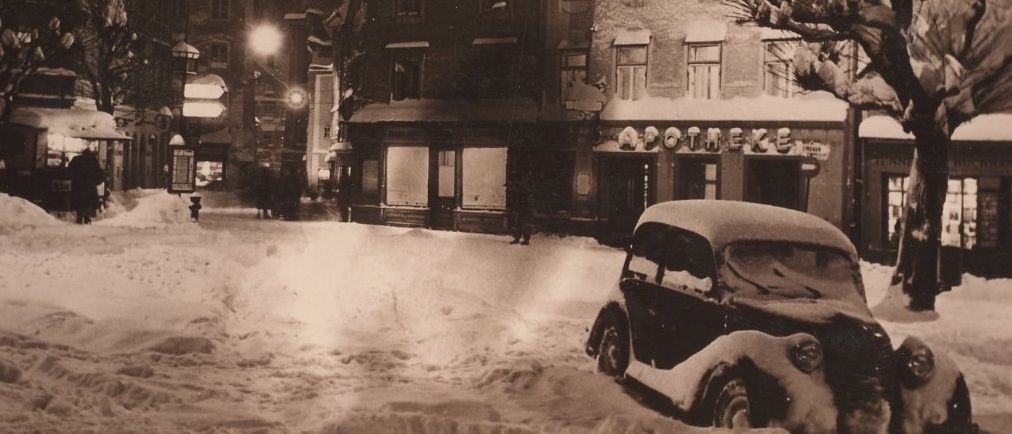 Eine verschneite Straße mit Gebäuden, einem geparkten Auto und Menschen, die gehen. Straßenlaternen und ein Schild für eine Apotheke.