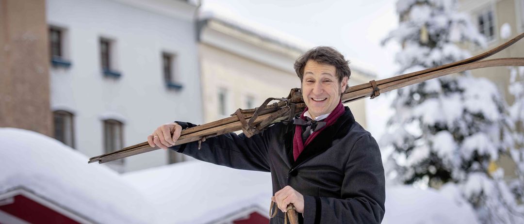 Ein Mann in Winterkleidung hält ein Paar Skier über seinem Kopf im Schnee und lächelt in die Kamera. Hinter ihm befinden sich Gebäude und ein verschneiter Baum.