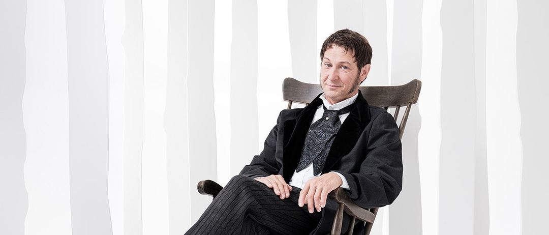 Ein Mann sitzt in einem hölzernen Schaukelstuhl in einer verschneiten Umgebung, trägt einen schwarzen Anzug und eine Krawatte. Er lächelt und schaut nach vorne, umgeben von Baumsilhouetten.