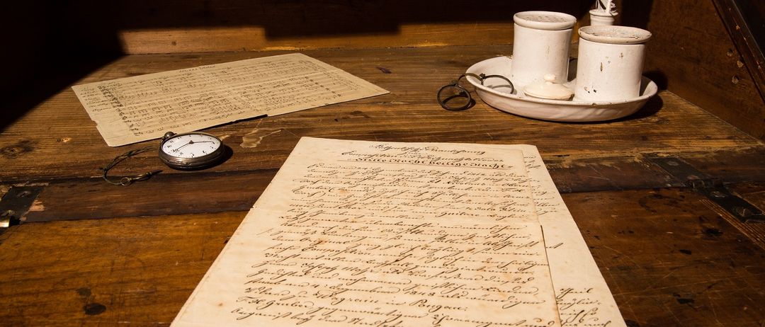 Ein alter Holzschreibtisch hat verschiedene Gegenstände darauf, einschließlich alter Dokumente, einer Feder, Tintenbechern und einer kleinen Uhr. Hinter dem Schreibtisch befinden sich Holzschubladen.