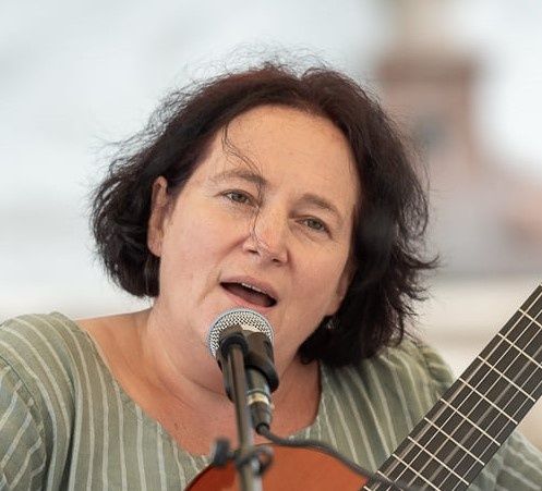 Eine Frau mit kurzen Haaren singt in ein Mikrofon und hält eine Gitarre. Sie trägt ein grün gestreiftes Hemd.