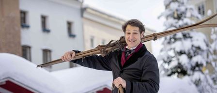 Ein Mann in einem schwarzen Mantel und Fliege hält ein Paar Skier und lächelt, steht im Schnee. Hinter ihm befindet sich ein schneebedecktes Gebäude und ein Weihnachtsbaum.