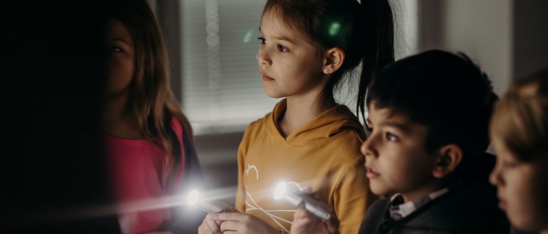 Drei Kinder spielen mit Taschenlampen in einem schwach beleuchteten Raum. Ein Mädchen in einer gelben Hoodie hält zwei Taschenlampen, während ein Junge in einem Anzug sie ansieht. Ein weiteres Mädchen in einem rosa Shirt steht hinter ihnen.