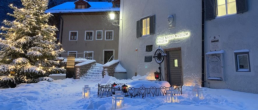 Eine Winterszene mit einem schneebedeckten Baum vor dem Stille Nacht Museum in Hallein, Österreich. Das Gebäude hat ein beleuchtetes Schild und mehrere Laternen.