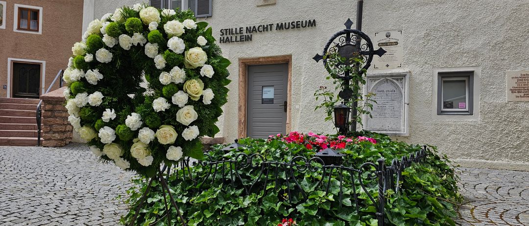 Ein Kranz aus weißen Rosen steht vor einem Gebäude mit der Aufschrift 'Stille Nacht Museum Hallein'. Das Gebäude hat eine Büste über der Tür und es gibt Pflanzen und Blumen drumherum.