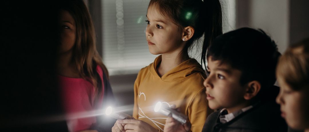 Drei Kinder stehen zusammen und halten Taschenlampen in der Hand. Sie scheinen an einer Aktivität beteiligt zu sein. Das Mädchen in der Mitte trägt eine gelbe Kapuzenjacke.