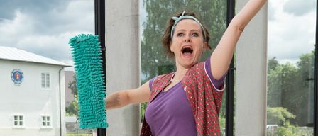 Eine Frau steht auf einem Hocker, hält ein Reinigungswerkzeug und trägt eine Reinigungskleidung. Sie scheint zu schreien und schaut nach oben. Hinter ihr befindet sich eine Glaswand mit Blick auf einen Fluss und ein Gebäude.