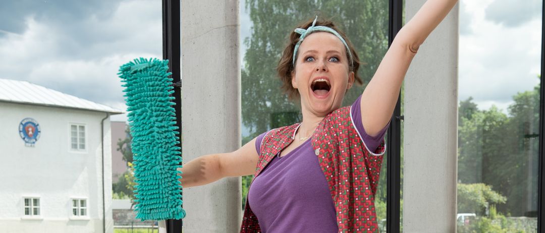 Eine Frau steht auf einem Hocker, hält ein Reinigungswerkzeug und trägt eine Reinigungskleidung. Sie scheint zu schreien und schaut nach oben. Hinter ihr befindet sich eine Glaswand mit Blick auf einen Fluss und ein Gebäude.