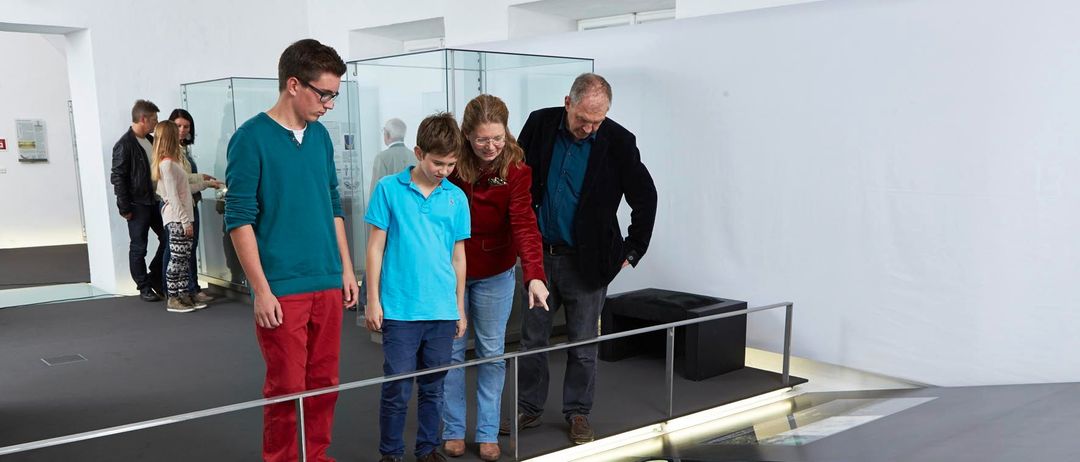 Vier Personen, vermutlich eine Familie, betrachten eine Ausstellung. Zwei von ihnen sind Kinder. Die Ausstellung ist in einer Glasvitrine ausgestellt.