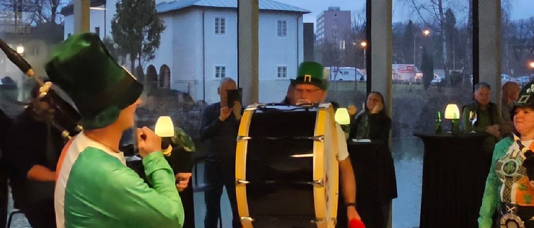 Eine Gruppe von Menschen in grünen und weißen Kostümen spielt drinnen Musikinstrumente. Ein Mann mit einer Dudelsack spielt vor einer großen Trommel. Draußen an den Fenstern gibt es Bäume und eine Kirche.