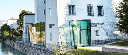 Ein weißes Gebäude mit Glasfenstern und einer grünen Tür steht neben einem Kanal, mit Bäumen und Bergen im Hintergrund.