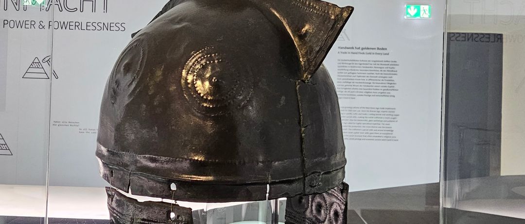 Ein antiker Helm wird in einer Glasvitrine ausgestellt. Der Helm ist aus Metall und hat komplizierte Muster. Er steht auf einer schwarzen Oberfläche mit einem weißen Schild davor.