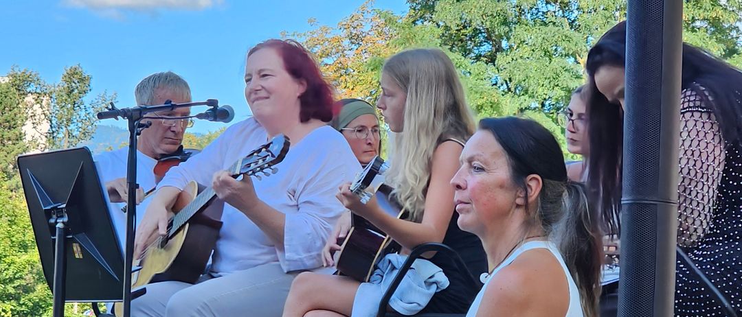 Eine Gruppe von Menschen ist auf einer Bühne, einige spielen Gitarre. Eine Frau vorn singt. Die Bühne ist draußen mit einem klaren blauen Himmel.