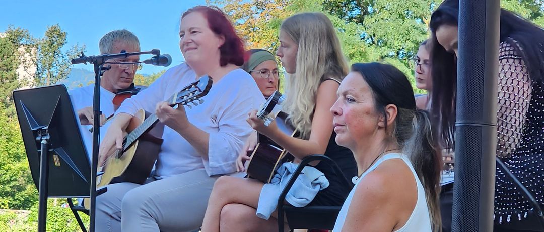 Eine Gruppe von Frauen und Männern spielt Gitarren und singt auf einer Bühne im Freien. Sie sitzen auf Stühlen, teilweise barfuß, mit einem Mikrofon und einem Notenständer.