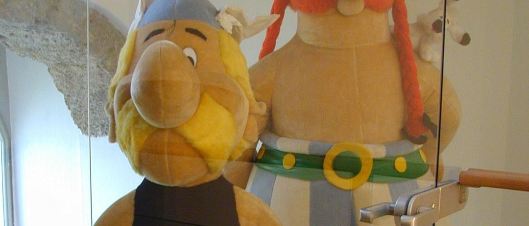 Zwei Plüschtiere von Charakteren sind in einem Glaskasten. Einer trägt einen Helm und hält ein gerolltes Objekt. Der andere trägt einen Gürtel mit Münzen.